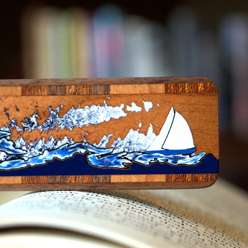 May include: A wooden bookmark featuring a blue and white sailboat design on a blue and white wave background. The bookmark is crafted from wood with a natural wood grain finish.