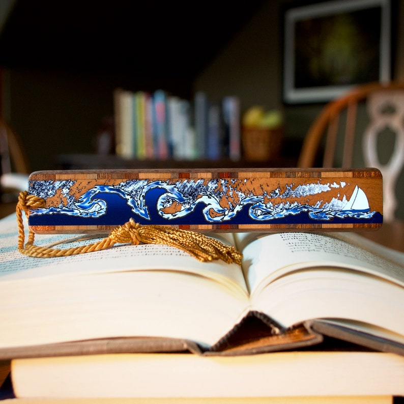 May include: A wooden bookmark featuring a blue and white wave design with a small white sailboat. The bookmark has a gold tassel.