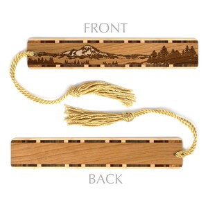 May include: Wooden bookmark with a laser-engraved image of a mountain range and trees. The bookmark has a brown wood grain and a gold tassel. The back of the bookmark is plain wood.