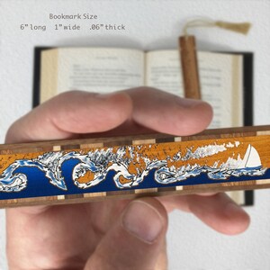 May include: Wooden bookmark featuring a blue and orange wave design with a white sailboat. The bookmark measures 6 inches long, 1 inch wide, and 0.06 inches thick.