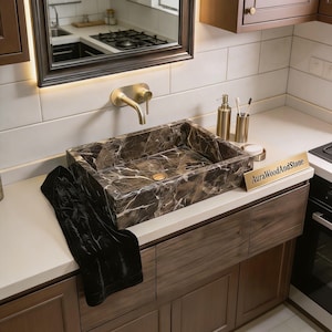 May include: A rectangular brown marble sink with white veining, set into a light-coloured countertop. A gold tap and matching soap dispenser, toothbrush holder, and small dish are visible. A black towel is draped over the counter. The text "AuraWoodAndStone" is on a small gold sign.