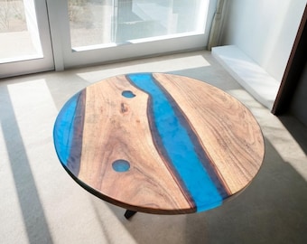 May include: Round wooden coffee table with a river of blue resin. The table has a natural wood grain finish with a glossy blue epoxy river design. The table is supported by three black legs. The table is approximately 91 cm in diameter.