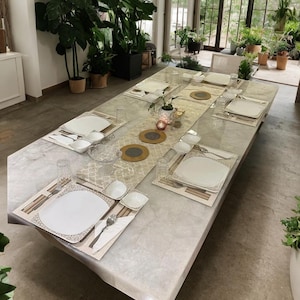 Könnte beinhalten: Ein Marmor-Esstisch, der für eine Mahlzeit gedeckt ist. Der Tisch ist mit weißen Tellern, Schalen, Besteck und Gläsern gedeckt. Ein dekorativer Tischläufer und Platzsets tragen zur Eleganz bei. Der Raum ist hell mit natürlichem Licht und Grün.