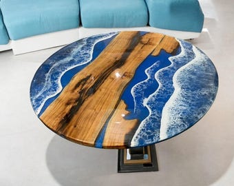 May include: Oval coffee table with a wooden top and a distinctive design. The table has a central wooden section with a natural grain, surrounded by blue resin that resembles ocean waves. It is supported by a black metal base.