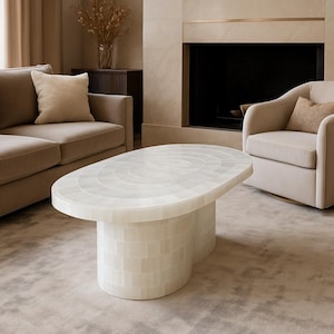 May include: Oval-shaped, off-white coffee table with a mosaic-like surface pattern. The base is a double-cylinder design. The table is in a living room setting with a sofa and armchair.