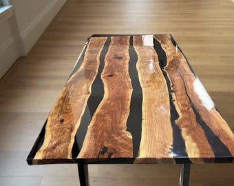 May include: A rectangular coffee table with a wooden top and black resin inlays. The wood has a natural grain pattern with shades of brown and tan. The table has a black metal base.