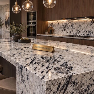 May include: A kitchen island featuring a white and black speckled granite countertop with a gold-toned triangular nameplate displaying the text AuraWoodAndStone. The island is paired with beige upholstered stools in a modern kitchen setting.