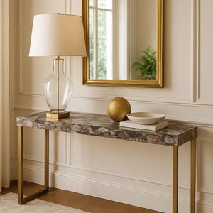 May include: A console table with a stone top and gold legs. A clear glass lamp with a cream shade sits on the table, along with a gold sphere and a white bowl. A gold-framed mirror hangs on the wall above the table. The table is approximately 120 cm long.