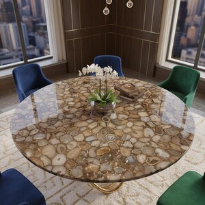 May include: A round dining table with a polished agate stone top, supported by a gold-colored base. The table is surrounded by four velvet chairs in blue and green. A chandelier hangs above the table, and a cityscape is visible through the window.