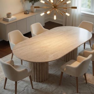 May include: A modern dining room set featuring a large oval table with a light-colored, textured surface. Six cream-colored chairs with wooden legs surround the table. A gold chandelier hangs above, and a neutral rug is beneath.