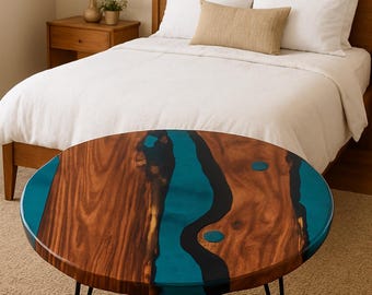 May include: A round coffee table featuring a wooden top with a teal epoxy resin river design. The table has a dark wood grain pattern and black hairpin legs. The table is in a bedroom setting.