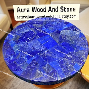 May include: A round table with a mosaic top made of deep blue stone pieces. The table has a dark edge and a gold-colored base. The text "Aura Wood And Stone" and a website address are visible.