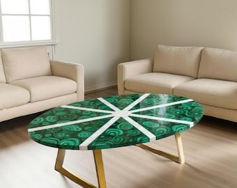 May include: Oval coffee table with a green malachite top and white starburst design. The table has gold-colored legs and is in a living room setting with beige sofas and a window.