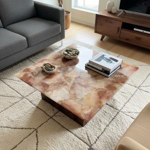May include: A square coffee table with a reddish-brown, patterned top and a dark brown base. The table sits on a cream-coloured rug with a geometric design. Two small bowls with stones and a stack of books are on the table.
