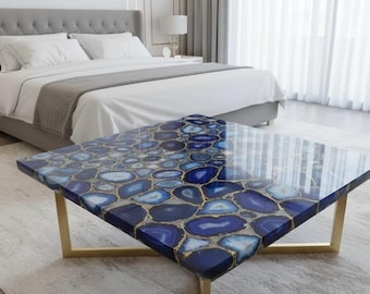May include: A square coffee table with a blue agate stone top and gold-colored metal legs. The agate design features shades of blue and white with gold accents. The table is in a bedroom setting.