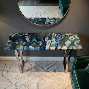 May include: A rectangular console table with a blue, white, and grey patterned top and polished chrome legs. The table is approximately 122 cm long and 41 cm wide. The table is in front of a grey wall and a large round mirror.