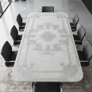 May include: A large, rectangular white table with ornate silver detailing, surrounded by eight gray upholstered chairs. The table is the focal point of a modern, well-lit room, suitable for dining or meetings.