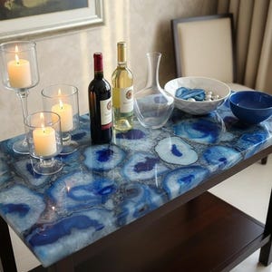 May include: A table with a blue agate stone top, featuring candles in glass holders, wine bottles, a decanter, and bowls. The table has a dark brown wooden frame. The table's top has a striking blue pattern.