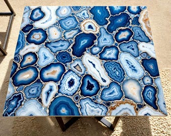 May include: A square table with a top made of blue and white agate slices. The agate pieces are arranged in a mosaic pattern, with gold-colored accents between the slices. The table has dark metal legs.