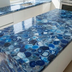 Puede incluir: Una isla de cocina y encimera con un llamativo diseño de ágata azul. Las superficies presentan un acabado pulido con una mezcla de patrones azules, blancos y grises. Los armarios son blancos y el suelo es de madera clara.