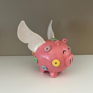 Puede incluir: Un cerdo de cerámica rosa con alas blancas y lunares de colores. El cerdo tiene una expresión feliz y está de pie en un estante blanco.
