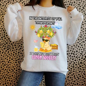 Könnte beinhalten: Ein weißes Langarm-Sweatshirt mit einer Grafik eines Enten-Motivs mit Sonnenbrille unter einem Zitronenbaum. Der Text lautet: IN THE INTEREST OF FULL TRANSPARENCY IT WAS NOT EASY PEASY LEMON SQUEEZY. Enthält eine Sonne, eine Wolke und einen Schmetterling.