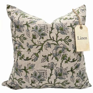 Puede incluir: Un cojín cuadrado de lino con un estampado floral en tonos verdes y azules sobre un fondo beige. El cojín tiene una etiqueta que dice "Linen". Un artículo de decoración para el hogar.