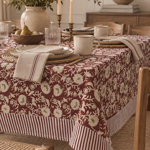 Puede incluir: Un mantel rojo oscuro con un patrón floral blanco y un borde a rayas. La mesa está puesta con tazas de cerámica, manteles individuales trenzados, servilletas beige con rayas rojas, vasos estriados y un cuenco de madera con peras.