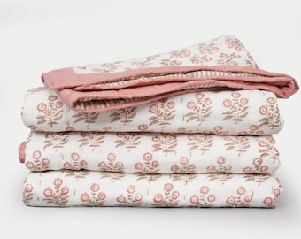 May include: A stack of four folded floral quilts. The quilts feature a white background with a repeating pattern of pink flowers and brown leaves. The top quilt has a pink border and a striped underside.