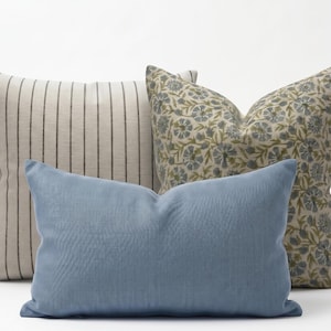 May include: Three decorative throw pillows. The front pillow is a solid light blue. Behind it is a pillow with a floral pattern in blue and green on a cream background. The third pillow has vertical stripes in a neutral color.