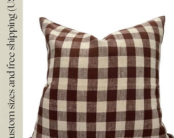 Puede incluir: Un cojín decorativo cuadrado con un patrón de cuadros marrones y crema. El cojín tiene un aspecto suave y texturizado y probablemente está hecho de tela. Es un artículo de decoración para el hogar.