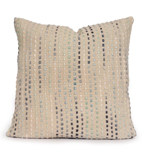 May include: A square throw pillow with a textured beige base fabric featuring vertical stripes of raised, stitched detailing in alternating shades of light blue, dark blue, and white, creating a subtle, repeating pattern across the entire surface.