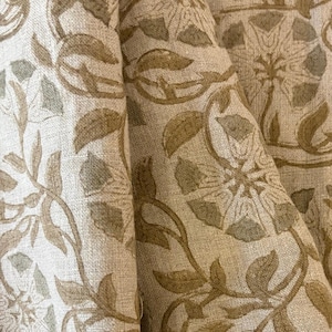 May include: Close-up of beige fabric with a repeating floral pattern in shades of brown and green. The design features stylized flowers and leaves, creating a natural, botanical theme. The fabric appears to be linen or a similar textured material.