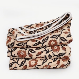May include: A stack of three folded floral patterned blankets. The blankets feature a beige background with brown flowers and black leaves. The edges are trimmed with white fabric.