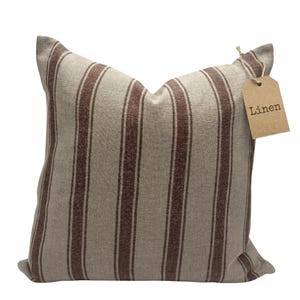 May include: A square throw pillow featuring a vertical striped pattern in shades of brown and beige. The pillow has a textured fabric surface and includes a brown paper tag attached with a string, with the word Linen printed on the tag.