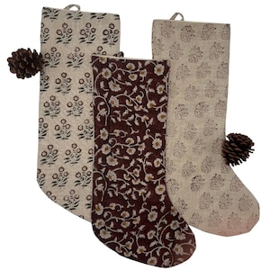 Peut inclure: Trois bas de Noël avec des motifs floraux. Deux sont beiges avec des motifs floraux bruns, et un est marron foncé avec des motifs floraux blancs. Chaque bas a une boucle pour accrocher et une pomme de pin.