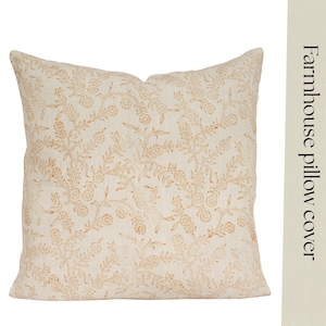 Puede incluir: Una funda de cojín cuadrada de tela color hueso con un patrón botánico repetitivo de color marrón claro que muestra pequeñas flores y enredaderas. El texto "Farmhouse pillow cover" aparece impreso verticalmente en negro a la derecha.