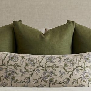 May include: A decorative pillow with a floral pattern in shades of green and blue, resting in front of three solid green pillows. The pillows are arranged on a neutral-colored surface, creating a layered and textured look.