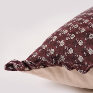 May include: A burgundy pillow with a white floral pattern and a ruffled edge. The pillow is made of a soft, textured fabric.