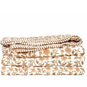 May include: A folded textile featuring a white base with a repeating pattern of tan floral motifs and green leaves. The top layer displays a contrasting design of thin, horizontal tan and white stripes, creating a layered appearance.
