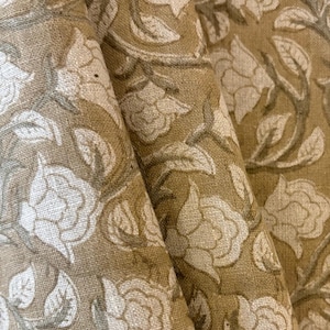May include: Close-up of folded fabric with a repeating floral pattern. The design features large, stylized white roses and leaves on a muted olive-green background. The fabric appears to be a natural fiber, possibly linen or cotton.