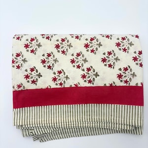 May include: Folded cream fabric featuring a floral design of red flowers and green leaves. A broad, solid red band is at the bottom, above a section with vertical green and white stripes. Likely a tablecloth or textile.