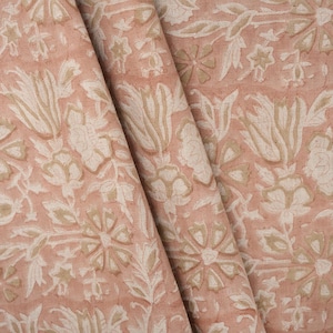 May include: Peach-colored fabric with a repeating floral pattern in off-white and tan. The design features stylized flowers and leaves, creating a textured, layered effect. The fabric is folded, showing the pattern's continuity.