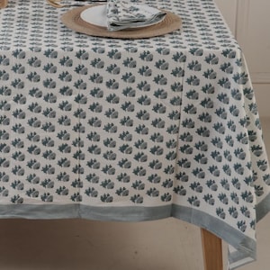 以下が含まれることがあります： 薄い青色の花柄が繰り返しプリントされた白いテーブルクロス。テーブルクロスは薄い青色の縁取りがあり、木製の脚のテーブルにかけられています。