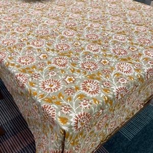 Może przedstawiać: Prostokątny obrus z powtarzającym się wzorem kwiatowym. Wzór przedstawia duże kwiaty w kolorach rdzy, bieli i musztardowej żółci na stonowanym zielonym tle. Obrus z tkaniny nadaje się na stół jadalny.