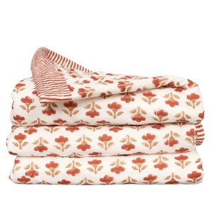 May include: A folded quilt featuring a cream-colored base with a repeating pattern of rust-colored flowers. The top edge of the quilt is folded over to reveal a rust and cream-colored striped border that contrasts with the floral print.