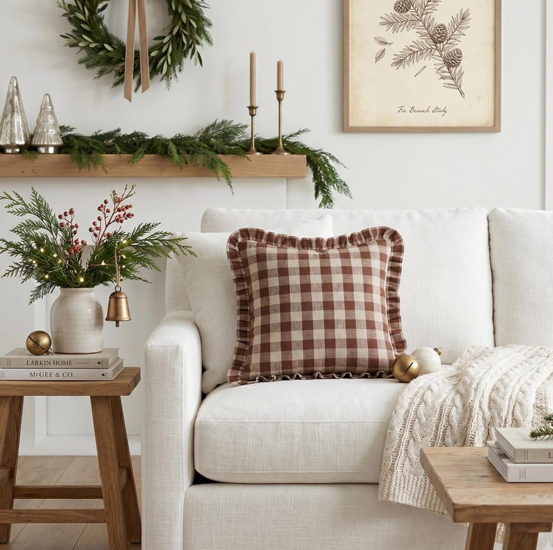 K&ouml;nnte beinhalten: Ein wei&szlig;es Sofa mit einem braun-cremefarbenen karierten Kissen mit R&uuml;schenkante. Ein h&ouml;lzerner Beistelltisch h&auml;lt eine Vase mit Gr&uuml;n und Beeren, eine Glocke und B&uuml;cher. Ein Kranz und ein gerahmter botanischer Druck h&auml;ngen an der Wand.