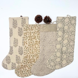 Peut inclure: Quatre bas de Noël avec des motifs floraux beiges, marron et gris. Les bas ont une boucle pour les suspendre et sont en tissu. Deux pommes de pin sont placées au-dessus des bas.