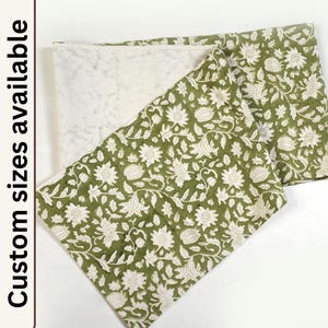 May include: A green fabric table runner with a white floral pattern. The runner is folded in half, showing the white fabric backing.