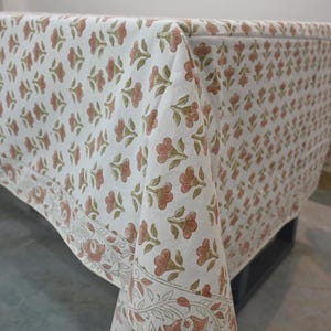 May include: A white tablecloth featuring a repeating pattern of small pink flowers with green stems and leaves. The fabric has a distinct border design along the edge, showcasing a continuous floral vine motif in matching pink and green tones.